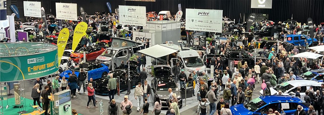 Gro&szlig;e Messehalle mit vielen Besuchern, zahlreichen Autos und St&auml;nden der Autohaus Peter Gruppe. Gelbe Peter-Fahnen und verschiedene Markenbereiche sind sichtbar.