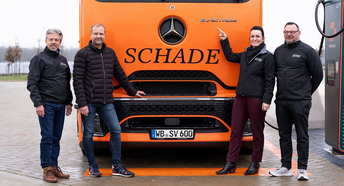 Vier Personen vor einem orangefarbenen Mercedes-Benz eActros-Lkw mit Schade-Logo. Eine Person zeigt auf das eActros-Emblem. Abgebildet von links nach rechts: Frank Hentschel (Verkaufsleiter Trucks, Autohaus Peter GmbH), Bj&ouml;rn Schade (Gesch&auml;ftsf&uuml;hrer Schade Logistics), Christina Dost (Gesch&auml;ftsf&uuml;hrerin Autohaus Peter GmbH), Enrico Dost (Serviceleiter).