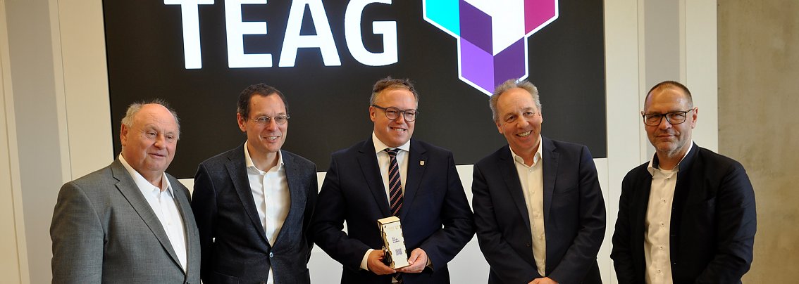 Das Bild zeigt die Jahreshauptversammlung des Landesverbandes des Kfz-Gewerbes Th&uuml;ringen, die in den R&auml;umen der Th&uuml;ringer Energie AG (TEAG) stattfand. F&uuml;nf M&auml;nner in Anz&uuml;gen stehen vor dem gro&szlig;en TEAG-Logo, der Mann in der Mitte h&auml;lt eine Auszeichnung oder einen Preis in den H&auml;nden. Alle l&auml;cheln in die Kamera, was auf eine positive, feierliche Atmosph&auml;re hinweist.