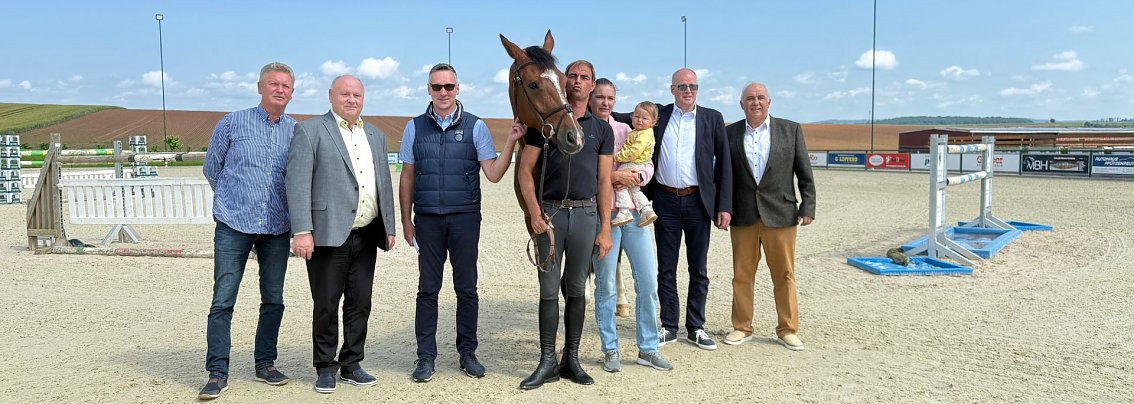 Pressekonferenz auf dem Reitplatz in Immenrode: Sieben erwachsene Personen stehen nebeneinander, darunter ein Reiter mit Pferd und eine Person mit Kind auf dem Arm. Im Hintergrund sind Hindernisse und die Reitplatzanlage zu sehen. Die Veranstaltung dient der offiziellen Vorstellung des Reitturniers Immenrode 2025.