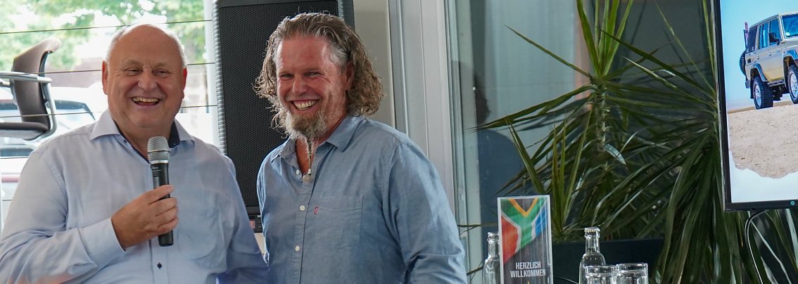 Afrikanischer Abend in Heiligenstadt: Helmut Peter und Abenteuer-Guide Hannu l&auml;cheln gemeinsam in die Kamera. Helmut Peter h&auml;lt ein Mikrofon, im Hintergrund sind ein Bildschirm mit Gel&auml;ndewagen und eine Pflanze zu sehen.