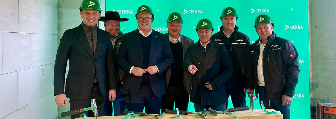 7 Herren mit gr&uuml;nen Dekra Helmen stehen vor einem gr&uuml;nen Dekra Banner und lachen in die Kamera