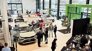 Im hellen Ausstellungsraum des �koda Autohauses steht ein neuer �koda Elroq mit ge&ouml;ffneten T&uuml;ren und Kofferraum. Mehrere G&auml;ste betrachten das Fahrzeug, einige unterhalten sich in kleinen Gruppen. Im Hintergrund sind weitere Autos sowie der Empfangsbereich mit der Beschilderung Information und Kasse zu sehen. Rechts am Rand ein Buffetbereich mit leeren Tassen und Tellern, daneben ein schwarzer Fotohintergrund.