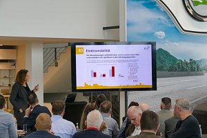 Teilnehmende verfolgen einen Vortrag im hellen �koda-Ausstellungsraum. Ein Bildschirm zeigt Diagramme, im Vordergrund Fr&uuml;hst&uuml;ckstische.