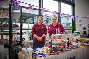 Zwei Frauen stehen hinter einem Kuchen- und Tortenstand im Autohaus. In einer Glasvitrine und auf Tischen sind Cupcakes und Torten ausgestellt. Rosa Flamingos dienen als Dekoration. Ein Banner mit dem Logo Happy Cakes h&auml;ngt &uuml;ber dem Stand.