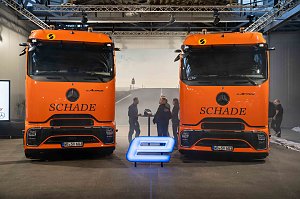 Zwei orangefarbene Mercedes-Benz eActros-Lkw mit Schade-Logo nebeneinander, davor ein blau leuchtendes E-Symbol. Personen im Hintergrund im Gespr&auml;ch.