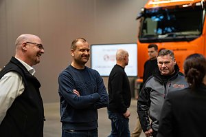Gruppe von Personen im Gespr&auml;ch vor einem orangefarbenen Mercedes-Benz eActros mit Schade-Logo. Im Vordergrund Bj&ouml;rn Schade im blauen Pullover.