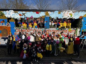 Gro&szlig;e Gruppe verkleideter Menschen posiert vor und auf einem bunt gestalteten Karnevalswagen mit M&auml;rchenmotiven, viele halten bunte Luftballons.