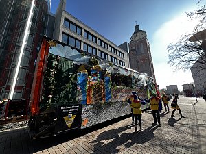 Seitliche Ansicht eines bunt dekorierten Karnevalswagens mit M&auml;rchenmotiven, gezogen von einem orangefarbenen LKW, im Hintergrund ein historisches Geb&auml;ude mit Uhrturm, mehrere als Narren verkleidete Personen laufen daneben.