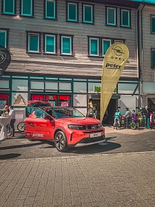 Roter Opel-Ausstellungswagen mit ge&ouml;ffneten T&uuml;ren neben einem gelben Opel-Banner, Passanten und Kinder im Hintergrund.
