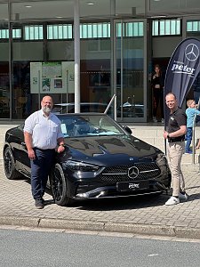 Zwei M&auml;nner stehen vor einem schwarzen Mercedes-Benz Coup&eacute; und l&auml;cheln in die Kamera. Rechts im Hintergrund ist eine Mercedes-Fahne, dahinter Glasfassade der Stadthalle.