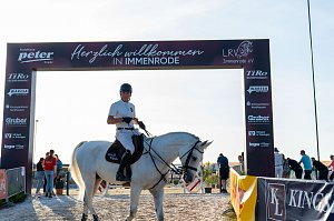Ein Reiter in wei&szlig;em T-Shirt und schwarzem Helm sitzt auf einem wei&szlig;en Pferd unter einem Begr&uuml;&szlig;ungsbogensorenn mit der Aufschrift �Herzlich willkommen in Immenrode�. Der Bogen ist mit Logos verschiedener Spo versehen. Im Hintergrund sind mehrere Personen und ein sonniger Himmel zu sehen.