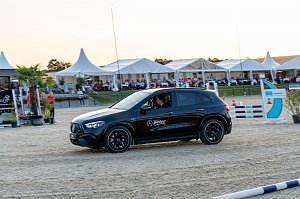 Ein schwarzer Mercedes-Benz SUV f&auml;hrt &uuml;ber den Sandplatz einer Reitanlage am Abend. Im Hintergrund sitzen Zuschauer unter wei&szlig;en Eventzelten und verfolgen das Turniergeschehen. [Mercedes-AMG GLA 35 4MATIC | Energieverbrauch kombiniert: 9,3-8,8 l/100 km | CO2-Emissionen kombiniert: 211-200 g/km | CO2-Klasse: G]