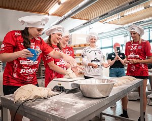 Spielerinnen des Thüringer HC in roten und weißen Trikots mit weißen Bäckermützen stehen in der Backstube der Bäckerei Helbing. Sie kneten und formen Teig, lachen und haben sichtlich Spaß. Im Vordergrund liegt Teig auf dem Tisch, daneben eine Schüssel mit Mehl. Eine Frau im Hintergrund filmt die Szene.