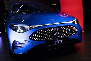 Nahaufnahme der Front eines blauen Mercedes CLA mit beleuchtetem K&uuml;hlergrill im Showroom. Ein rotes Eventbanner mit Mercedes-Logo ist im Hintergrund zu sehen.