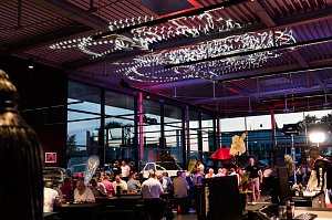 Viele G&auml;ste sitzen an langen Tischen in der beleuchteten Eventhalle. An der Decke sind dekorative Lichtinstallationen angebracht.