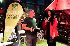 Christina Dost im roten Blazer moderiert neben einem Mann im gr&uuml;nen T-Shirt vor einem gelben Banner mit der Aufschrift M&ouml;bius � Gr&uuml;n macht gl&uuml;cklich!. Hinter ihnen ein schwarzer Mercedes-Camper.