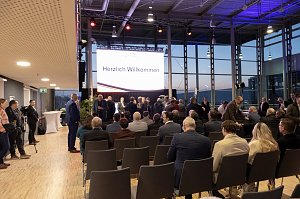 Publikum in einer Messehalle vor einer B&uuml;hne mit gro&szlig;er Leinwand Herzlich Willkommen, G&auml;ste im Anzug nehmen Platz.