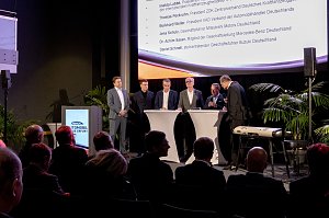 Podiumsrunde auf der B&uuml;hne der Automobil Messe Erfurt mit mehreren M&auml;nnern im Anzug, Publikum im Vordergrund und Teilnehmerliste auf der Leinwand.