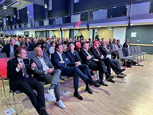 Publikum sitzt in Reihen in einer Messehalle, mehrere M&auml;nner im Anzug in der ersten Reihe.