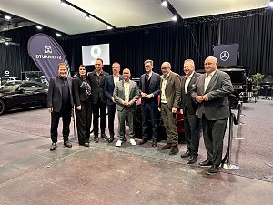 Gruppenfoto von neun Personen auf einer Automobilmesse, im Hintergrund Ausstellungsfahrzeuge und Markenbanner.