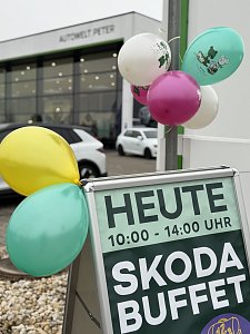 Werbeschild mit Text Heute 10:00�14:00 Uhr Skoda Buffet, dekoriert mit bunten Luftballons vor einem Autohaus.