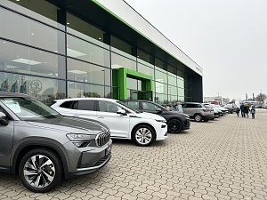 Reihe geparkter Autos vor einem modernen Autohaus mit gro&szlig;er Glasfront und gr&uuml;nem Eingang, Menschen gehen &uuml;ber den Parkplatz.