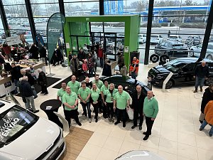 Mitarbeitergruppe in gr&uuml;nen Poloshirts steht im Autohaus zwischen ausgestellten Skoda-Fahrzeugen, im Hintergrund viele Besucher am Eingang.