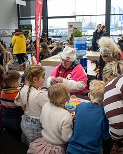 Kinder basteln an einem Tisch, betreut von einer Frau im Pinken Pullover; im Hintergrund Besucher in einem Autohaus.