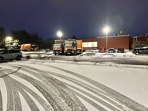 Winterliche Szene auf einem Betriebshof bei Nacht mit mehreren orangefarbenen M&uuml;llfahrzeugen und verschneitem Parkplatz.