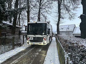 M&uuml;llfahrzeug f&auml;hrt auf einer schmalen, verschneiten Stra&szlig;e zwischen H&auml;usern und B&auml;umen.