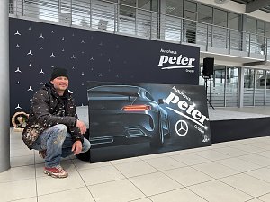 Alex Sitt hockt neben dem fertigen Graffiti-Bild. Auf dem Bild ist ein Pkw von hinten zu sehen, daneben ist das Logo der Autohaus Peter Gruppe.