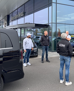 Vier M&auml;nner stehen vor der modernen Glasfassade des Mercedes-Benz Autohauses Peter. Links ragt das Heck einer schwarzen V-Klasse ins Bild.