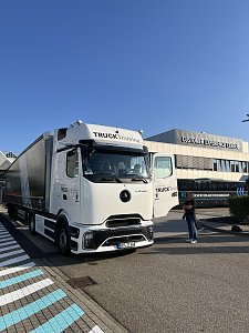 Ein wei&szlig;er Mercedes-Benz Lkw mit Sattelauflieger steht bei strahlendem Sonnenschein vor einem Mercedes-Benz Geb&auml;ude. Die Fahrert&uuml;r der Zugmaschine ist ge&ouml;ffnet, daneben steht eine Person.