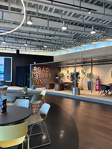 Ein gro&szlig;z&uuml;giger Loungebereich mit dunklem Holzboden und moderner Deckenbeleuchtung zeigt im Hintergrund eine Sitzecke mit der Aufschrift ROAD STARS INN. Im Vordergrund stehen mehrere runde Tische mit gelben, grauen und wei&szlig;en St&uuml;hlen.