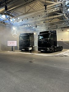 Zwei baugleiche, gl&auml;nzende schwarze Mercedes-Benz Lkw stehen nebeneinander in einer hell erleuchteten &Uuml;bergabehalle. An der linken Wand wird ein leuchtender Mercedes-Stern projiziert. Daneben steht ein Bildschirm mit dem Logo von Ehrlich Logistics.