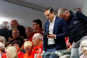 Ein Mann im dunkelblauen Anzug blickt auf sein Smartphone, w&auml;hrend Zuschauer in roten Trikots auf der Trib&uuml;ne der Handballhalle sitzen.